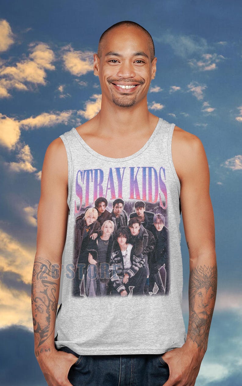 May include: A light gray tank top featuring a group of people with the text "STRAY KIDS" in a purple and pink gradient. The tank top is worn by a person with a blue sky background.