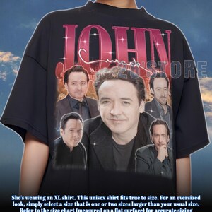 May include: Black unisex t-shirt featuring a collage of images of John Cusack, with the name "JOHN" in large, pink, sparkling letters. The shirt includes the text "23 STORE".