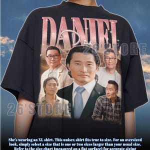 May include: Black unisex t-shirt featuring a portrait of a man with the name "DANIEL" in a pink, rose gold font. The shirt also has the text "26'STORE" in white. The shirt is an XL size.