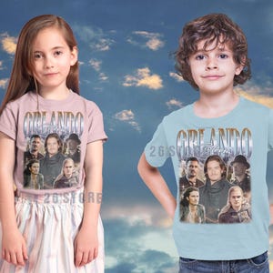 May include: Two children wearing t-shirts with the word "Orlando" in a vintage style font. The shirts feature a collage of images of people. The girl is wearing a pink shirt and the boy is wearing a light blue shirt. The text "26'S STORE" is at the bottom.