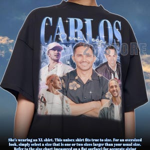 Op de afbeelding: Zwart unisex T-shirt met de naam "CARLOS" in een blauw, gestileerd lettertype. Het shirt toont meerdere afbeeldingen van een man. De tekst "256TORE" is zichtbaar. Het shirt is maat XL.
