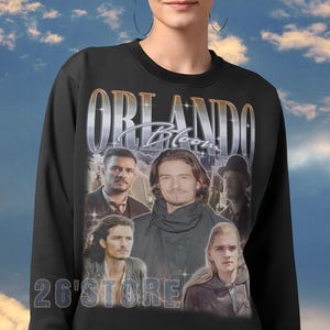 May include: Black crewneck sweatshirt featuring a collage of images of Orlando Bloom, with the name "ORLANDO Bloom" in a silver font. The text "26'STORE" is printed below the collage.