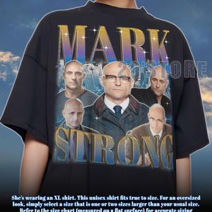 May include: Black unisex t-shirt featuring a collage of images and the text "MARK STRONG" in a gold and blue gradient font. The shirt has a crew neck and short sleeves.