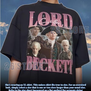 Puede incluir: Camiseta unisex negra con un diseño gráfico de estilo vintage. El diseño presenta el texto "LORD BECKETT" en rosa y blanco, junto con retratos de un hombre con vestimenta histórica. La camiseta también tiene el texto "26'STORE".