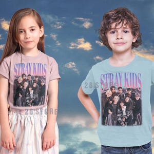 May include: Two children wearing t-shirts with the text "STRAY KIDS" in a gradient font. The shirts feature a group photo of the band. The girl is wearing a pink shirt and the boy is wearing a light blue shirt.