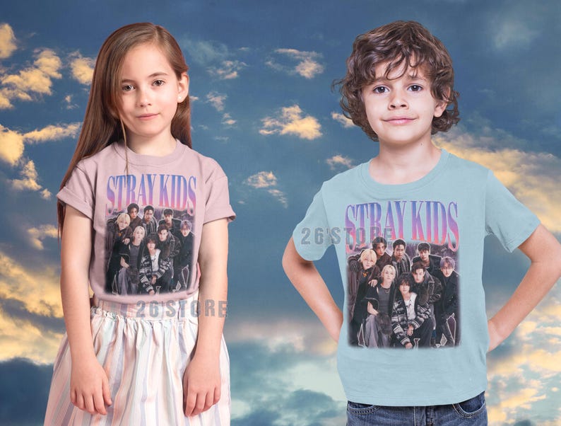 May include: Two children wearing t-shirts with the text "STRAY KIDS" in a gradient font. The shirts feature a group photo of the band. The girl is wearing a pink shirt and the boy is wearing a light blue shirt.