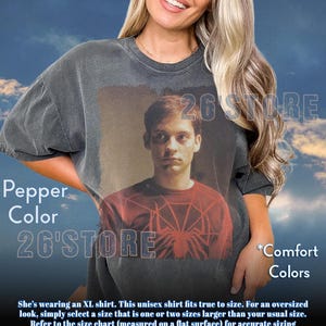 May include: A charcoal grey oversized t-shirt featuring a graphic print of a person wearing a red spider-web patterned shirt. The t-shirt has a crew neckline and short sleeves. The text 26'STORE is visible on the shirt and in the background.
