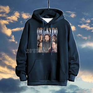 May include: Navy blue hooded sweatshirt featuring a graphic of Orlando Bloom and the text "ORLANDO Bloom". The sweatshirt has a front pocket and a drawstring hood. The text "26'Store" is printed at the bottom.