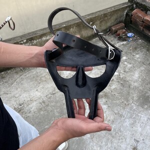 May include: A black metal mask with a distinctive, stylized design, featuring a nose and mouth area, and two vertical bars extending downwards. A black leather strap with a silver buckle is attached to the mask.