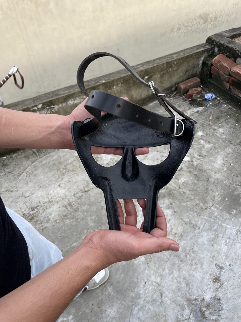May include: A black metal mask with a distinctive, stylized design, featuring a nose and mouth area, and two vertical bars extending downwards. A black leather strap with a silver buckle is attached to the mask.