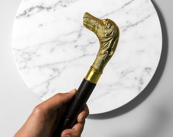 May include: A decorative walking cane with a brass wolf head handle and a black wooden shaft. The handle is detailed with intricate carvings. The cane is held against a white marble background.