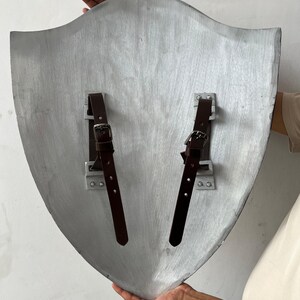 May include: A silver-colored shield with a curved top and pointed bottom. Two brown leather straps with buckles are attached to the back with metal brackets. The shield is held up by a person's hands.