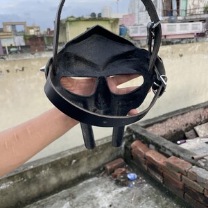 May include: Black leather mask with eye and mouth openings, featuring a strap for wearing. The mask has a distinctive, angular design and is held up by a strap. The mask is made of leather and has a dark, matte finish.