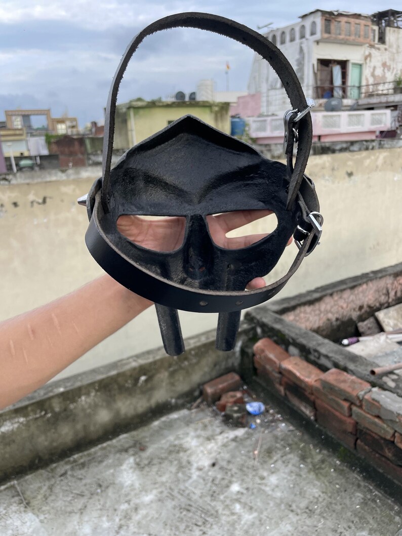 May include: Black leather mask with eye and mouth openings, featuring a strap for wearing. The mask has a distinctive, angular design and is held up by a strap. The mask is made of leather and has a dark, matte finish.