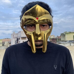 May include: A gold-colored mask with a distinctive design, featuring a prominent face and a red jewel. The mask is worn over the face. The person is wearing a black shirt.