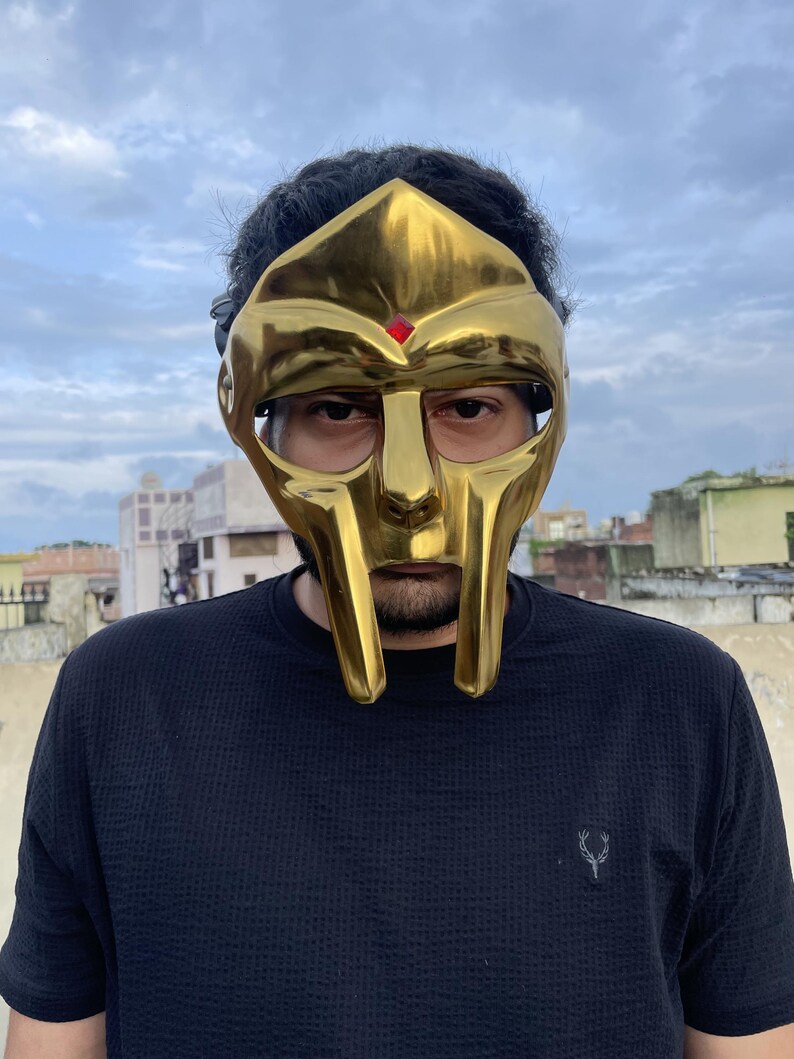 May include: A gold-colored mask with a distinctive design, featuring a prominent face and a red jewel. The mask is worn over the face. The person is wearing a black shirt.