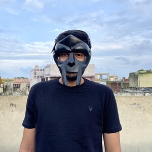 May include: A black metal mask with a distinctive design, featuring a prominent nose and mouth area, and eye openings. The mask is worn by a person wearing a black t-shirt. The background shows a cloudy sky and buildings.
