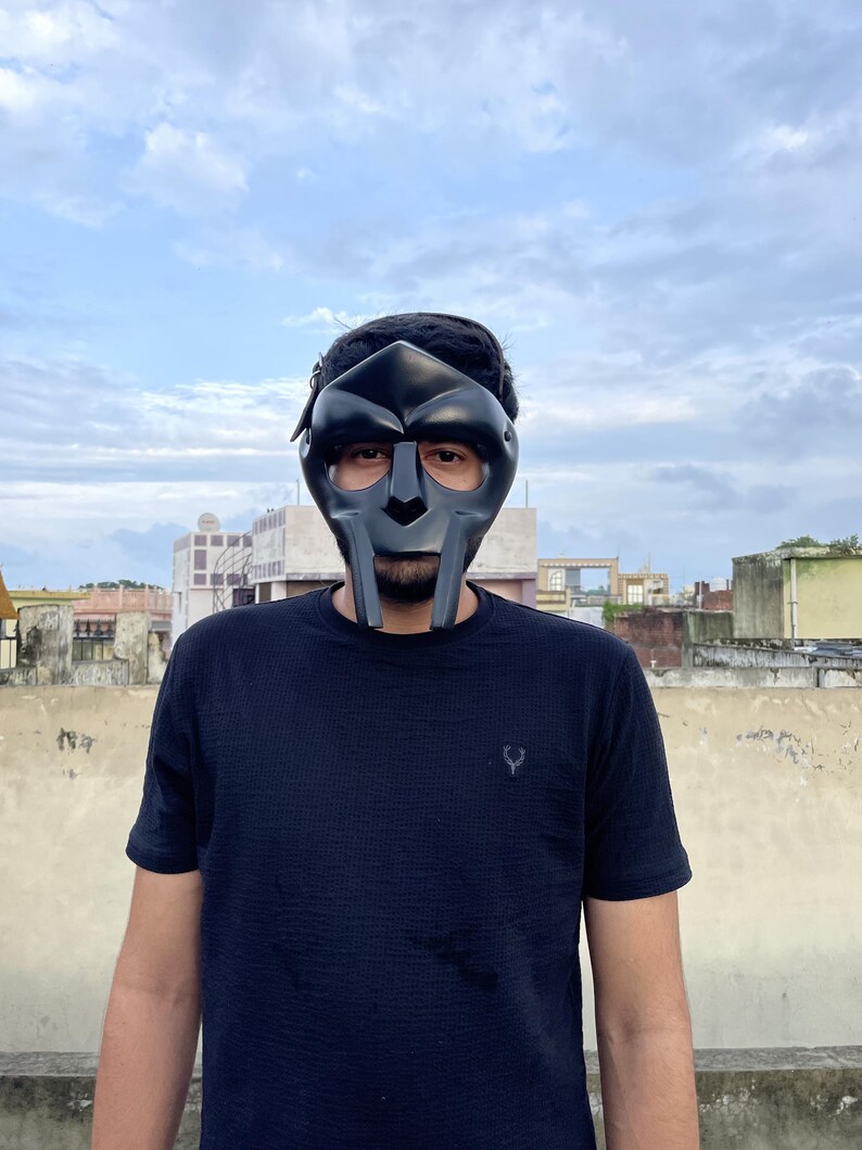 May include: A black metal mask with a distinctive design, featuring a prominent nose and mouth area, and eye openings. The mask is worn by a person wearing a black t-shirt. The background shows a cloudy sky and buildings.