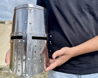 May include: A polished, silver-colored knight's helmet with a flat top and riveted construction. The helmet features horizontal eye slits and numerous small ventilation holes. The design is reminiscent of medieval armor.