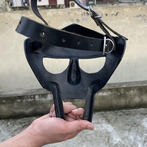 May include: A black metal mask with a distinctive design, featuring a nose and mouth area, and two vertical bars extending downwards. It has a black leather strap with a metal buckle for securing the mask.