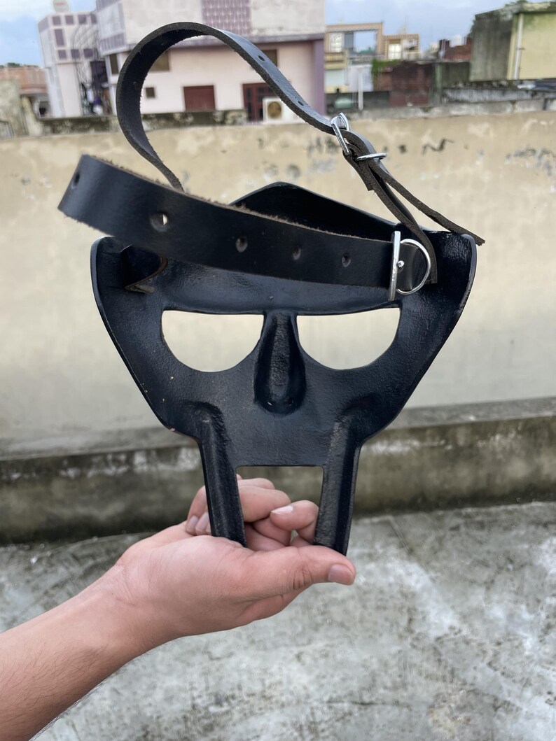May include: A black metal mask with a distinctive design, featuring a nose and mouth area, and two vertical bars extending downwards. It has a black leather strap with a metal buckle for securing the mask.