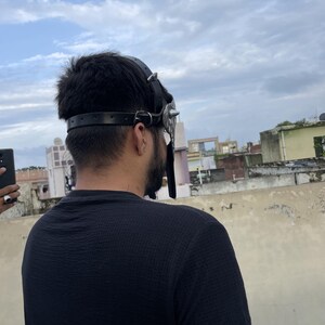 May include: A black leather mask with silver spikes and studs. The mask has a strap that goes around the head and is worn by a person. The person is wearing a black shirt and is standing outside.