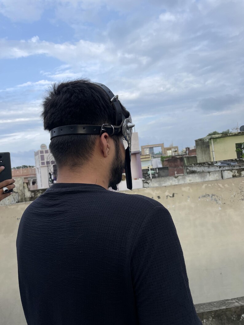 May include: A black leather mask with silver spikes and studs. The mask has a strap that goes around the head and is worn by a person. The person is wearing a black shirt and is standing outside.