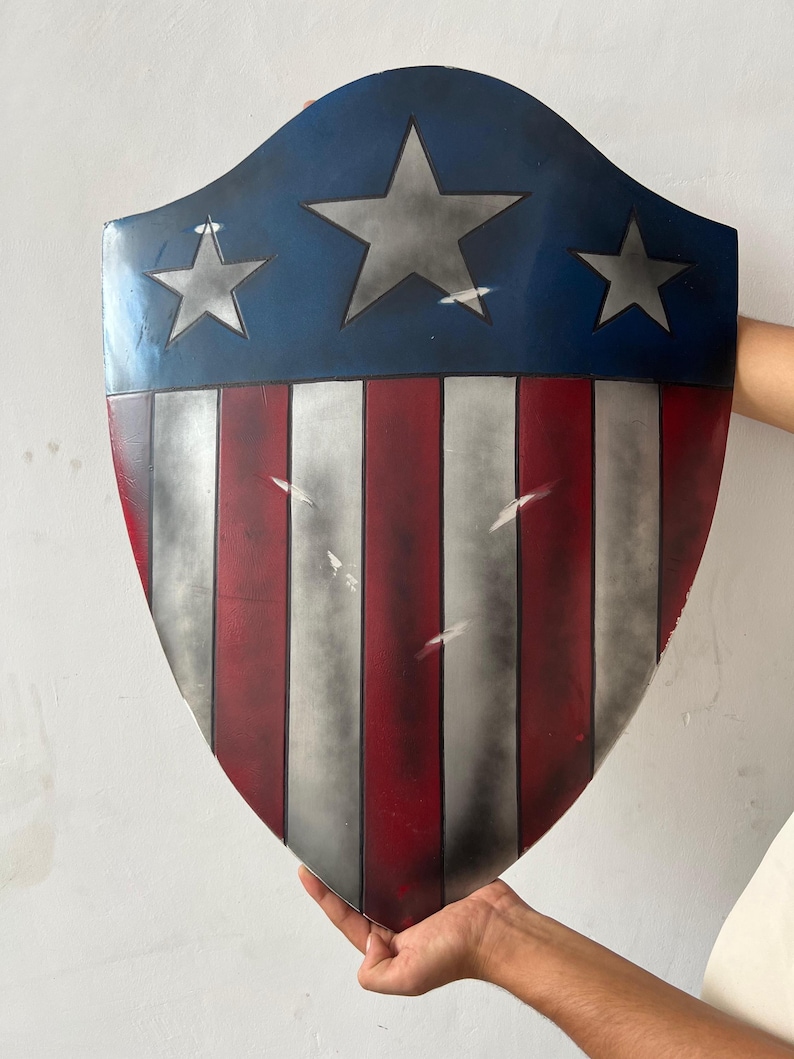 May include: A shield with a blue top section featuring three gray stars. The lower portion has alternating red and white vertical stripes. The shield is held up against a white background.