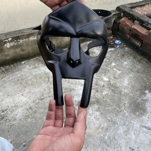 May include: A black mask with a distinctive, angular design. The mask features a prominent nose and a wide, rectangular mouth area. It has eye openings and a strap for securing it. The mask is held up by a person's hands.