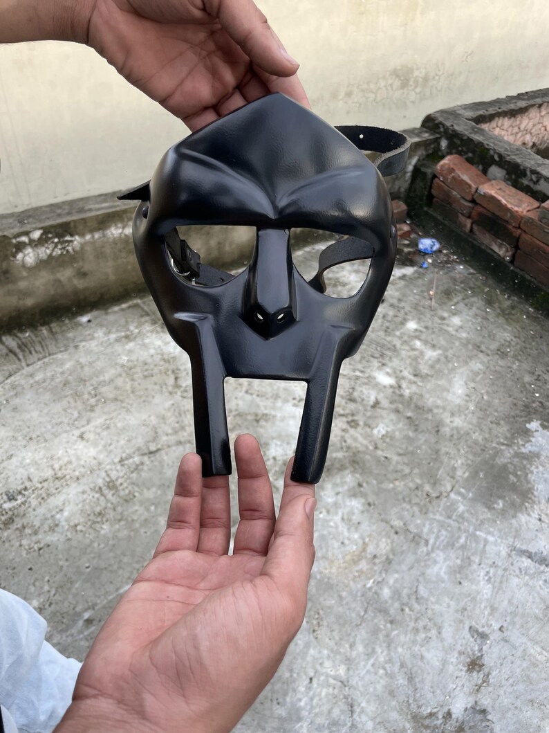 May include: A black mask with a distinctive, angular design. The mask features a prominent nose and a wide, rectangular mouth area. It has eye openings and a strap for securing it. The mask is held up by a person's hands.