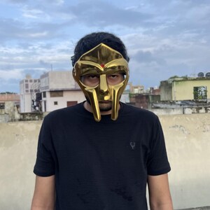 May include: A gold-colored mask with a distinctive design, featuring a prominent face shape and eye openings. The mask has a smooth, reflective surface and a small red accent. The person is wearing a black t-shirt.