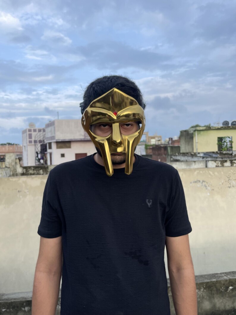 May include: A gold-colored mask with a distinctive design, featuring a prominent face shape and eye openings. The mask has a smooth, reflective surface and a small red accent. The person is wearing a black t-shirt.