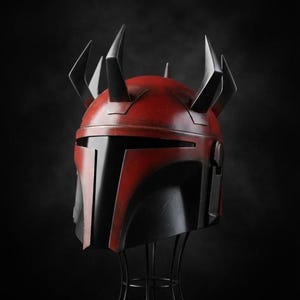 May include: A red and black helmet with multiple black spikes on top, displayed on a black wire stand. The helmet has a distinctive angular design, with a red top and black faceplate. The background is a solid black.