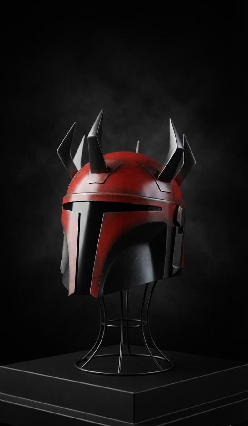 May include: A red and black helmet with multiple black spikes on top, displayed on a black wire stand. The helmet has a distinctive angular design, with a red top and black faceplate. The background is a solid black.