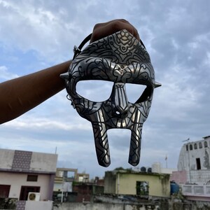 May include: A silver-colored mask with intricate engraved designs and pointed studs. The mask has large eye openings and a distinctive, stylized face shape. A black strap is attached to the mask.