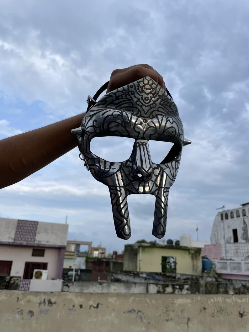 May include: A silver-colored mask with intricate engraved designs and pointed studs. The mask has large eye openings and a distinctive, stylized face shape. A black strap is attached to the mask.