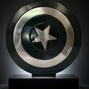 May include: A black and silver shield with a five-pointed star in the center. The shield has a circular design with a silver band and is displayed on a black rectangular base. The shield is approximately 24 inches in diameter.