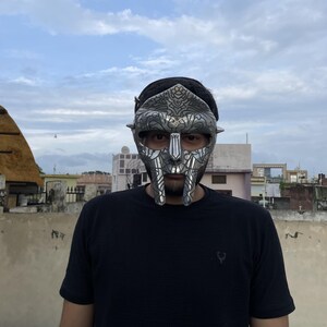 May include: A silver-colored mask with intricate details, worn by a person. The mask covers the upper face, featuring a stylized design with a prominent nose and mouth area. The person is wearing a black t-shirt.