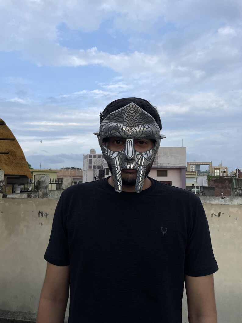 May include: A silver-colored mask with intricate details, worn by a person. The mask covers the upper face, featuring a stylized design with a prominent nose and mouth area. The person is wearing a black t-shirt.