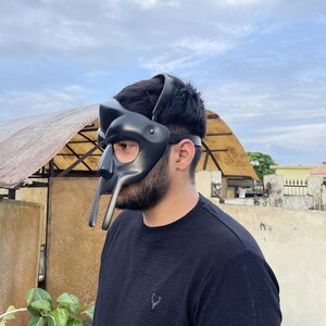 May include: A black mask with a unique design, featuring a prominent nose piece and extended lower elements. The mask is worn on the head with a strap. The person is wearing a black shirt.