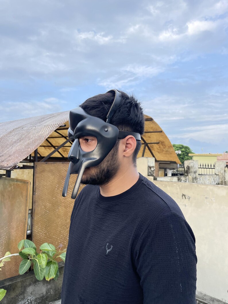 May include: A black mask with a unique design, featuring a prominent nose piece and extended lower elements. The mask is worn on the head with a strap. The person is wearing a black shirt.