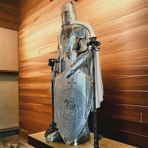 Może przedstawiać: Srebrna statua rycerza z białą peleryną i tarczą z krzyżem i napisem "XPS TI MILITIA DEI" w okrągłym wzorze. Statua stoi na drewnianej powierzchni.
