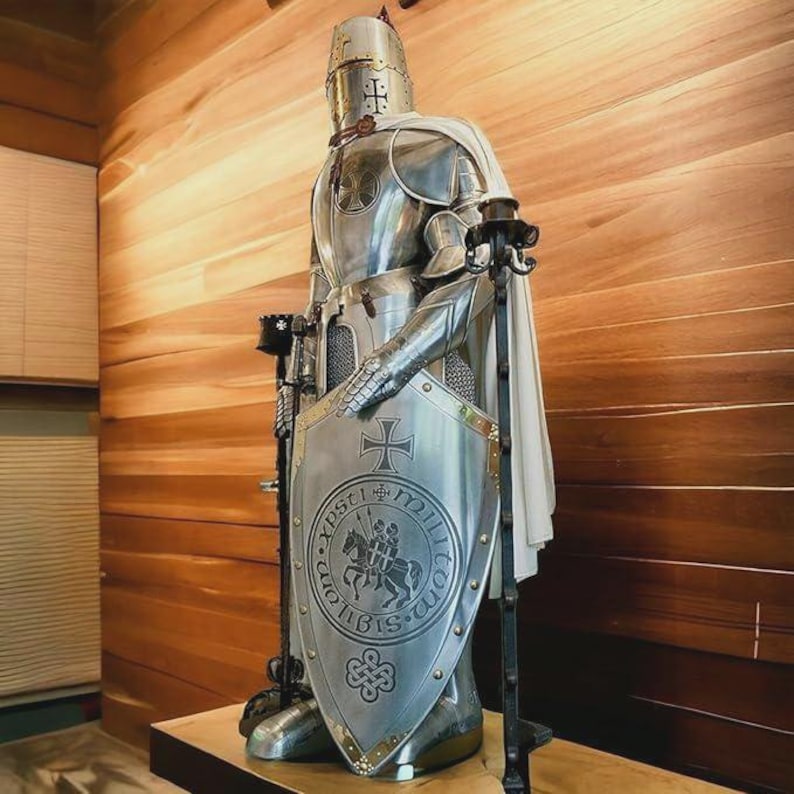 Może przedstawiać: Srebrna statua rycerza z białą peleryną i tarczą z krzyżem i napisem "XPS TI MILITIA DEI" w okrągłym wzorze. Statua stoi na drewnianej powierzchni.