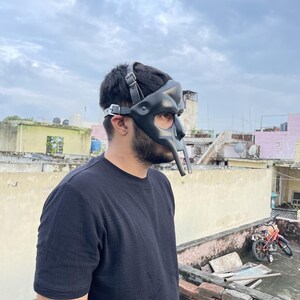 May include: A black leather mask with a unique, stylized design, featuring a prominent nose piece and adjustable straps. The mask is worn by a person wearing a black shirt. The background shows a cloudy sky and rooftops.