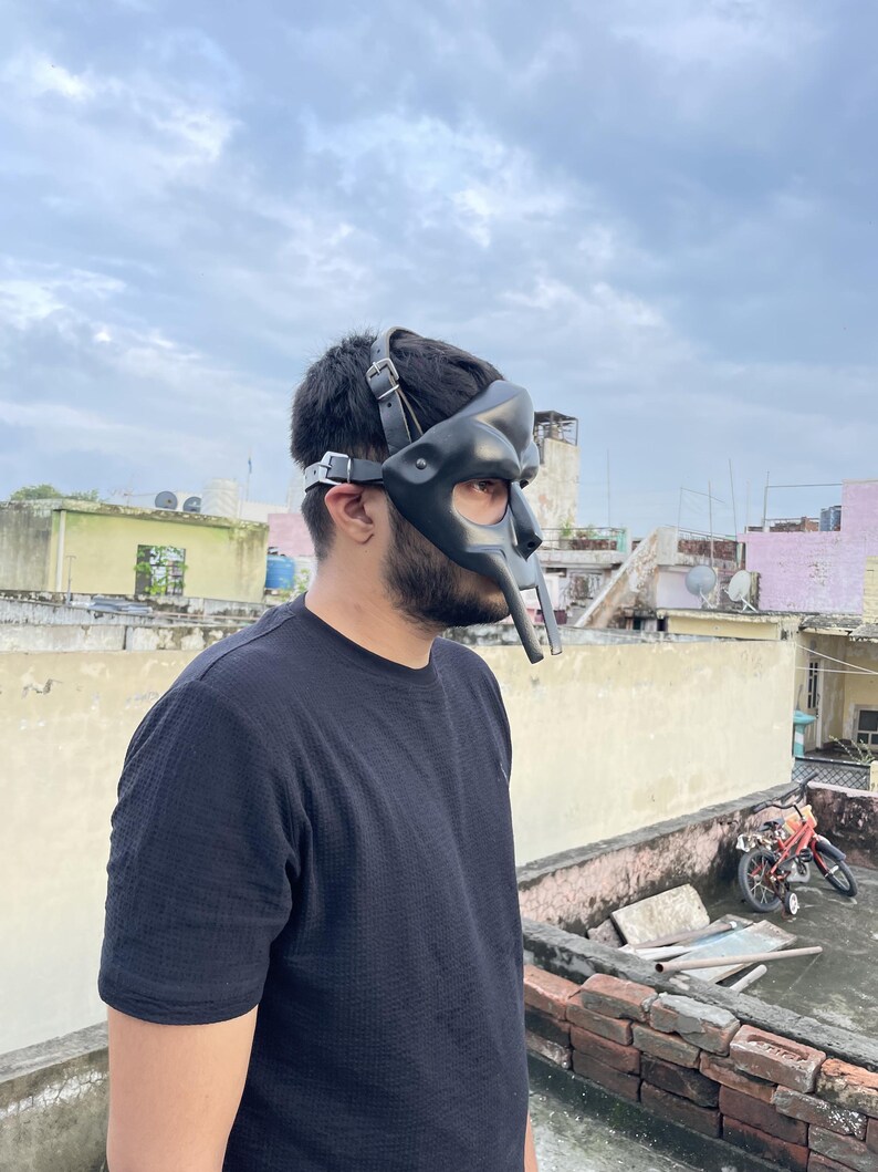 May include: A black leather mask with a unique, stylized design, featuring a prominent nose piece and adjustable straps. The mask is worn by a person wearing a black shirt. The background shows a cloudy sky and rooftops.