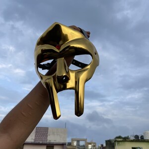 May include: A gold-colored mask with a distinctive design, featuring elongated facial features and eye openings. The mask is held up against a cloudy sky, showcasing its reflective surface and unique shape. The mask appears to be made of metal.