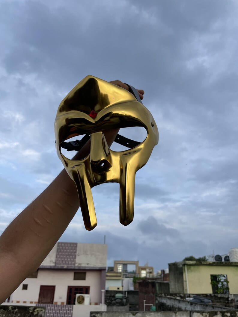 May include: A gold-colored mask with a distinctive design, featuring elongated facial features and eye openings. The mask is held up against a cloudy sky, showcasing its reflective surface and unique shape. The mask appears to be made of metal.