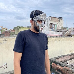 May include: A silver and gray gladiator mask with intricate designs. The mask covers the face and has a head strap. The person is wearing a black t-shirt. The background is a cloudy sky and rooftops.