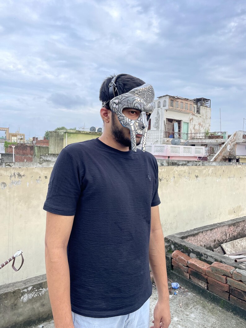 May include: A silver and gray gladiator mask with intricate designs. The mask covers the face and has a head strap. The person is wearing a black t-shirt. The background is a cloudy sky and rooftops.