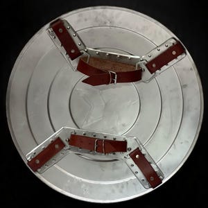 May include: A silver-colored, circular shield with a star emblem in the center. The shield has brown leather straps and metal brackets for securing the shield to the arm. The shield is a replica of a superhero's shield.